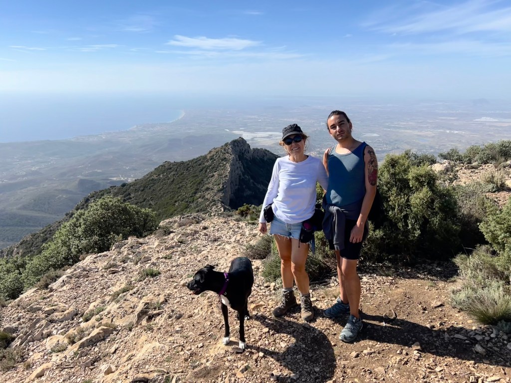 Ana y Miguel posan junto a la perra Xena en una montaña de Alicante, con el paisaje y el mar al fondo.